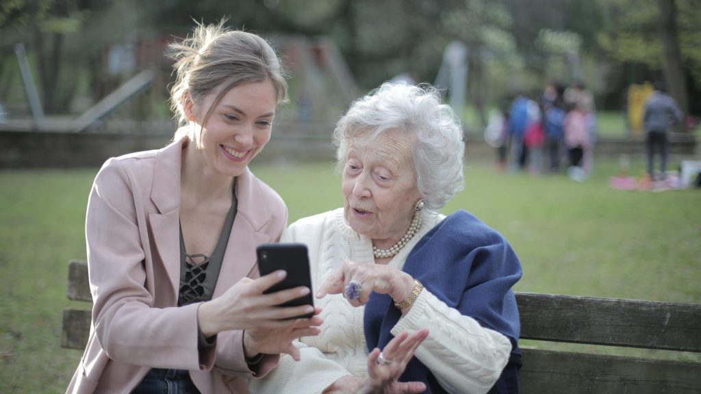 a young woman sitting on a bench with an older woman, looking at a phone as an example of a hearing technology partner