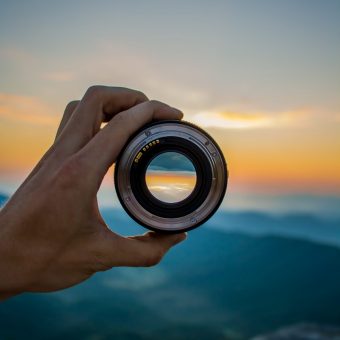 hand holding a camera lens with focused image against a blurry sunset background